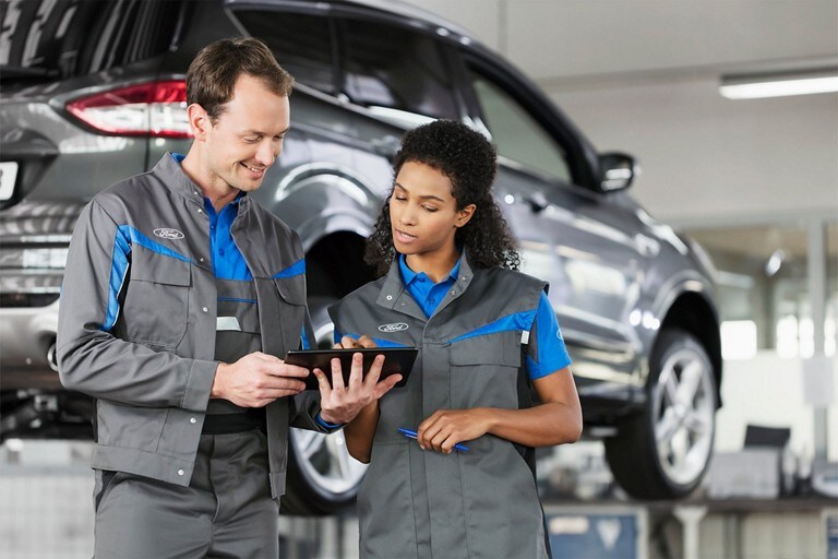 Two Ford employees in front of a vehicle on a car lift looking at a tablet.